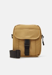 Tan canvas schoudertas met een klep sluiting, zwarte gesp en verstelbare schouderband. Kenmerkt zich door een gladde textuur en een compacte rechthoekige vorm.