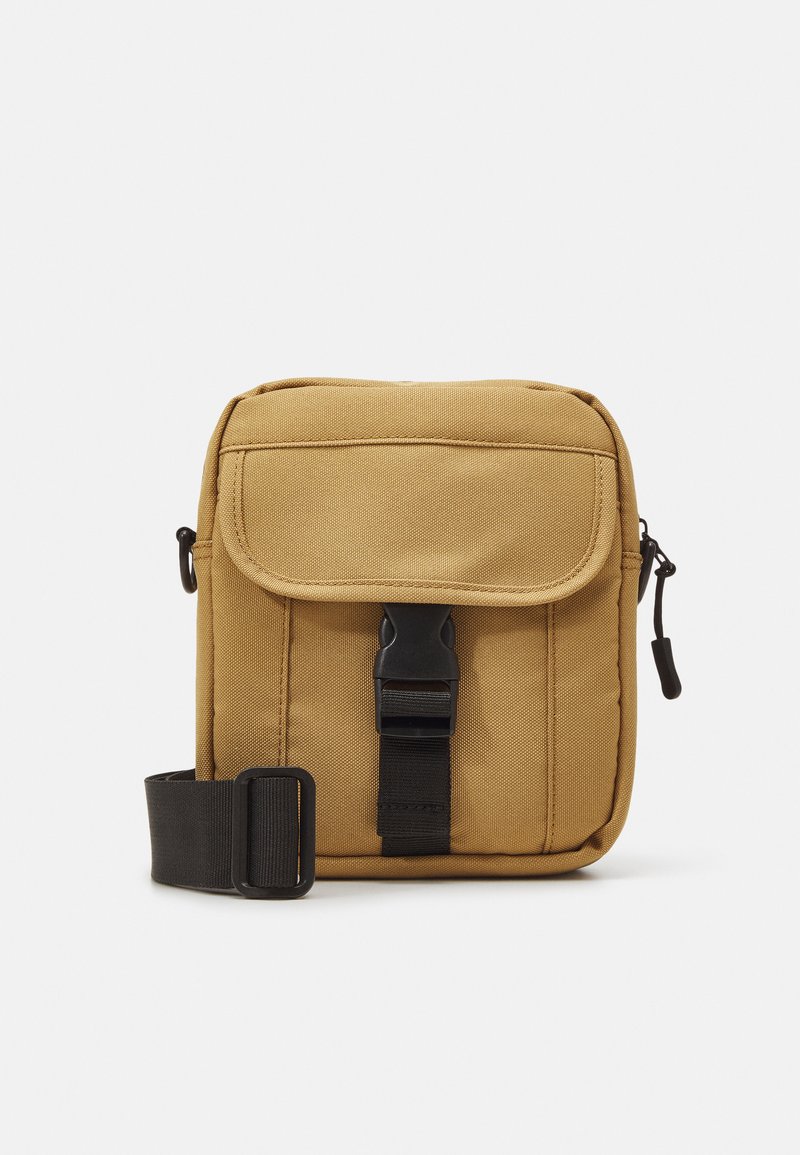 Tan canvas schoudertas met een klep sluiting, zwarte gesp en verstelbare schouderband. Kenmerkt zich door een gladde textuur en een compacte rechthoekige vorm.