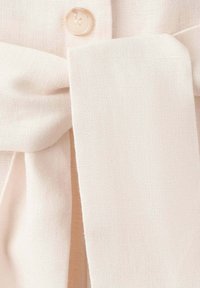 Gros plan d'une ceinture en tissu crème nouée avec un bouton assorti sur un fond de vêtement en lin texturé.