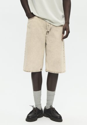 Muž v béžových riflových kraťasech po kolena, šedých ponožkách a černých šněrovacích botách, stojící s jednou rukou v kapse.