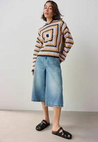Kolorowy sweter na szydełku z geometrycznym wzorem, sparowany z jasnoniebieskimi spodniami culotte i czarnymi sandałami na paski. Miękkie tekstury i luźny krój.