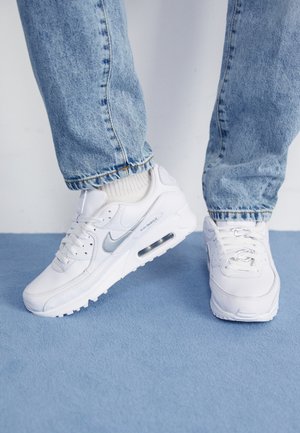 Osoba nosząca białe buty sportowe Nike Air Max z białymi skarpetkami i jasnoniebieskimi jeansami stoi na niebieskim dywanowym podłożu.