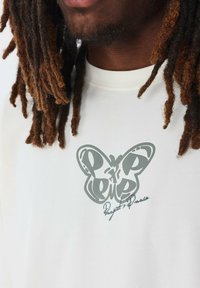 Camiseta de cuello redondo color crema con un gráfico de mariposa gris y texto manuscrito abajo, hecha de una tela suave con una textura lisa.