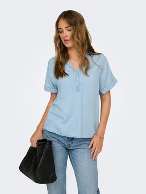 Donna che indossa una camicetta azzurra a maniche corte e jeans, tiene una borsa nera con una mano, su uno sfondo chiaro e uniforme.