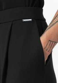 Black trousers featuring a textured fabric, wide waistband with pleats, and a silver hardware accent displaying the brand name.
