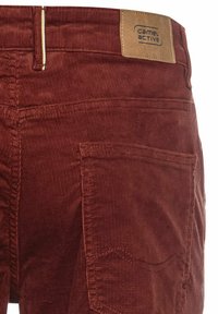 Rote Corduroyhosen in Rostfarbe mit strukturiertem Oberflächen, Stickdetails an den hinteren Taschen und einem Lederlogo-Label am Bund.