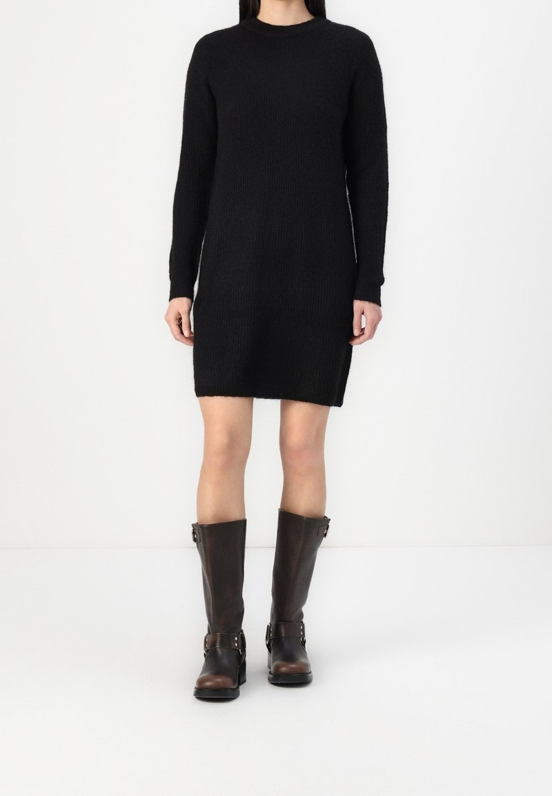 Femme portant une robe en maille noire à manches longues associée à des bottes en cuir marron foncé mi-mollet sur fond blanc.
