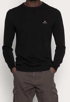 Homme portant un pull noir à col ras du cou avec un petit logo blanc GANT sur la poitrine et un pantalon cargo marron, les mains dans les poches.
