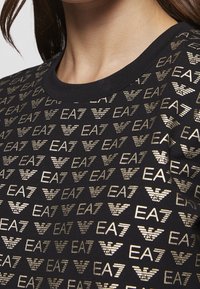 Svart t-shirt med rund halsringning, prydd med ett upprepat mönster av gyllene EA7-logotyper och en mjuk bomullstextur.