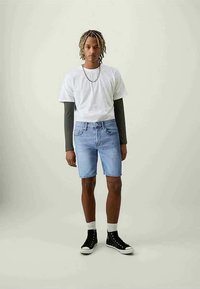Shorts di jeans azzurri con una vestibilità rilassata, abbinati a una t-shirt bianca e maniche lunghe grigie. Un paio di sneakers alte nere completano il look.
