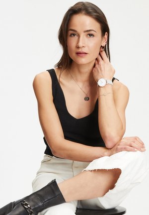 Model, der ein schwarzes geripptes Tanktop, weiße, ausgefranste Jeans und schwarze Stiefel mit Reißverschluss trägt. Zubehör beinhaltet eine silberne Uhr und mehrlagige Halsketten.
