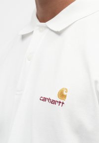 Witte poloshirt gemaakt van textiel met textuur, voorzien van een kraag en drie knopen, met een gouden en kastanjebruin geborduurd Carhartt-logo.