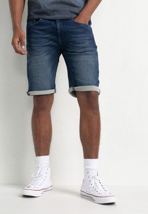 Jeansowe szorty w ciemnoniebieskim kolorze z podwiniętymi mankietami, z przednimi kieszeniami i dopasowanym krojem. Noszone z białymi wysokimi trampkami i skarpetkami.