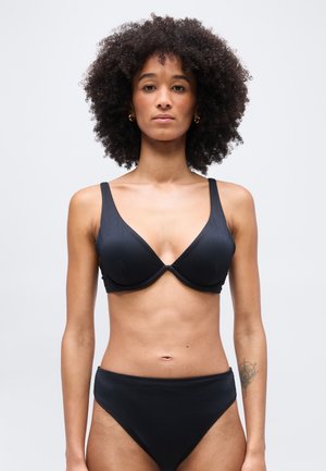 Žena s kučeravými vlasmi, oblečená v čiernom rebrovanom podprsenke a zodpovedajúcich nohavičkách s vysokým pásom, stojaca pred jednoduchým svetlošedým pozadím.