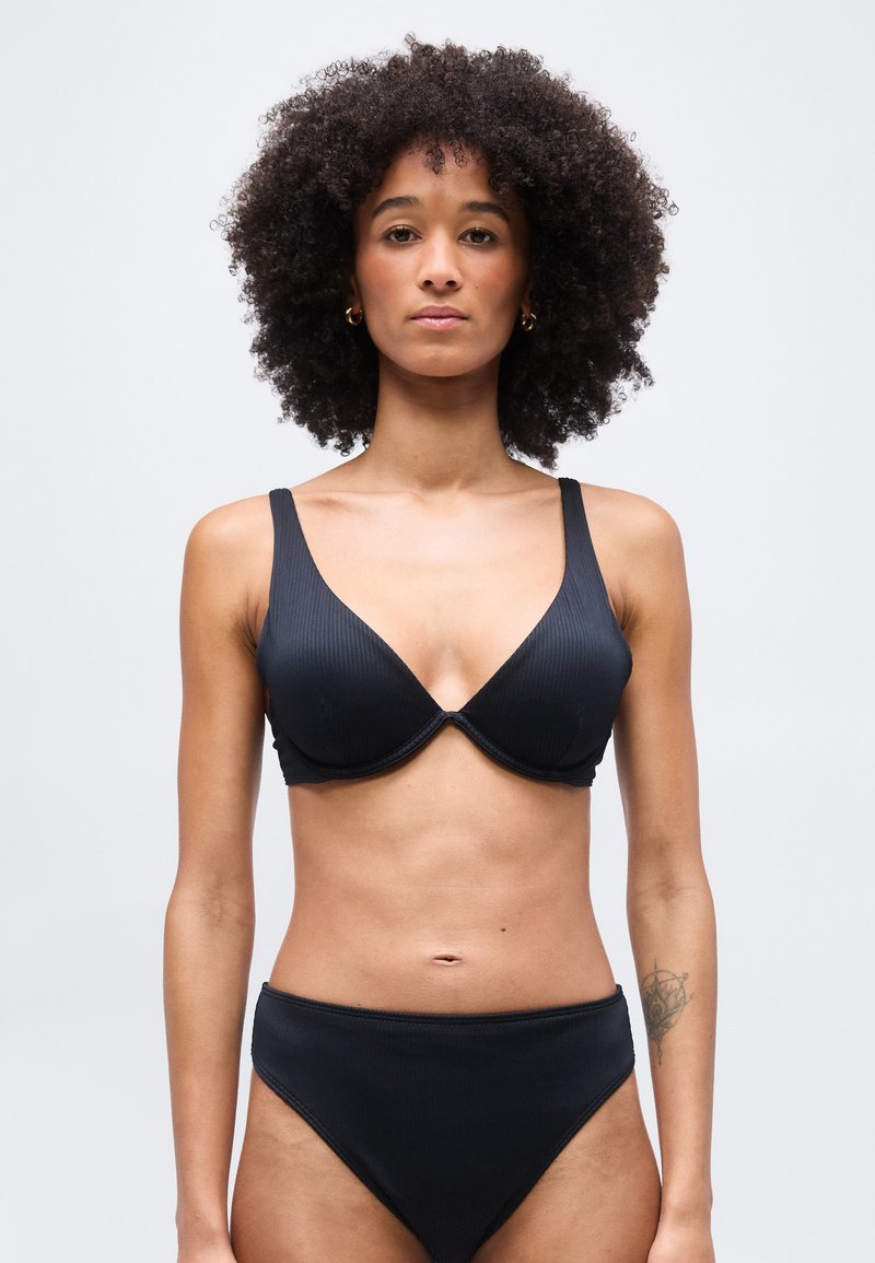 Γυναίκα με σγουρά μαλλιά που φοράει μαύρο ριμπ σουτιέν και αντίστοιχο σλιπ με ψηλή μέση, στέκεται μπροστά σε απλό ανοιχτό γκρι φόντο.