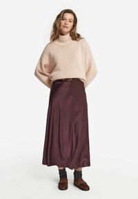 Przesadnie duży, jasnoróżowy sweter z dzianiny z prążkowanym golfem, w połączeniu z bordową satynową spódnicą midi, brązowymi mokasynami oraz ciemnoszarymi skarpetkami.