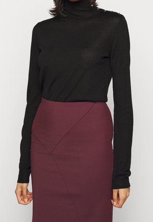 Kobieta ubrana w czarny golf z długim rękawem, wsunięty do wysokiej taliowanej, ciemnofioletowej spódnicy ołówkowej z subtelnymi przeszyciami.