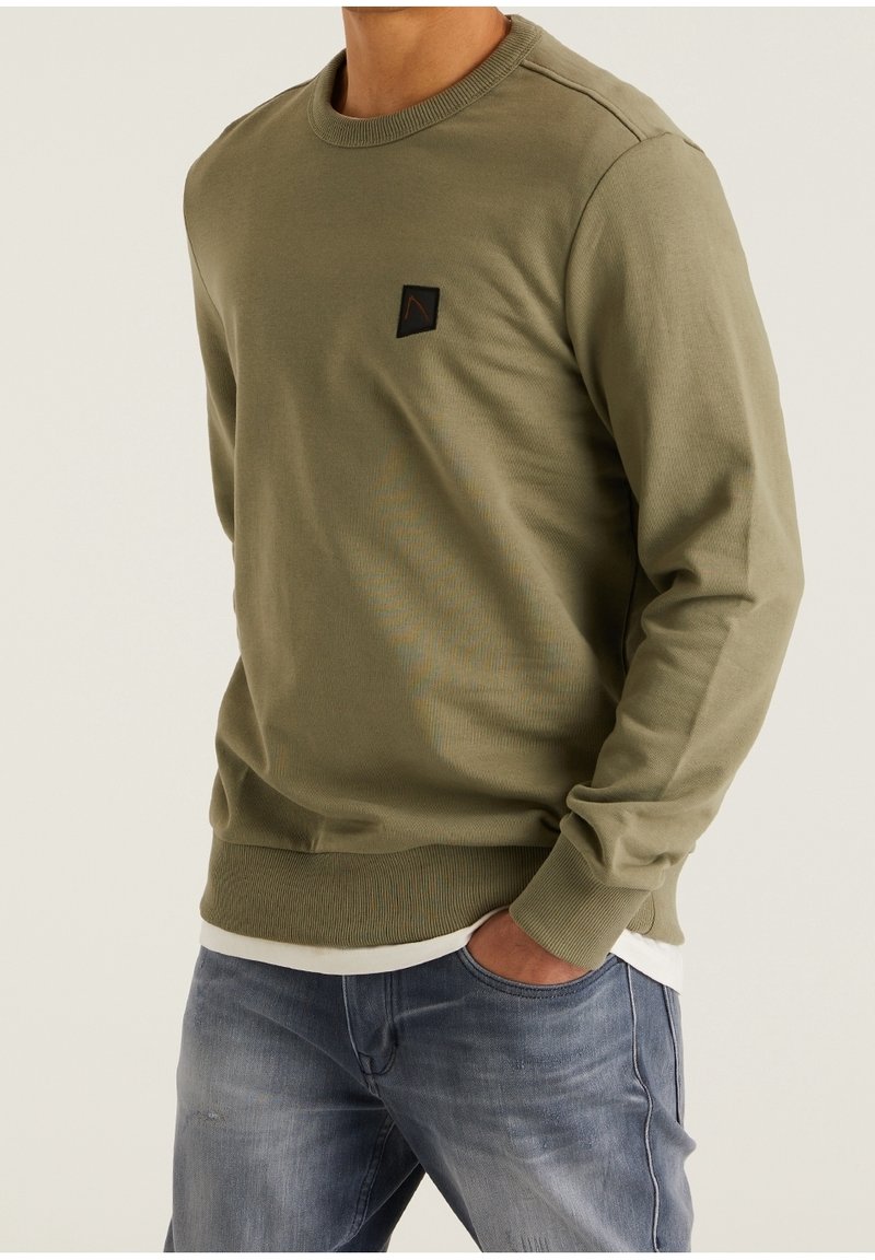 Personne portant un pull ras du cou vert olive avec un petit écusson noir et rouge sur la poitrine, associé à un jean bleu délavé et un sous-vêtement blanc.