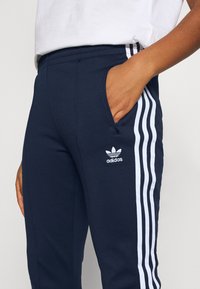 Mörkblå Adidas-joggingbyxor med vita ränder längs sidorna, med en ficka, elastisk midja och en liten logotyp nära höften.