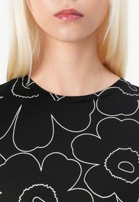 Svart topp med blomsterdesign i hvitt, med rund halslinje og glatt tekstur. Designet fremhever store, stiliserte blomster.