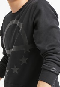 Svart sweatshirt med en stor grå logotyp som har en cirkel och stjärnor. Tillverkad av ett slätt tyg med ribbade ärmslut och en Nike Air-detalj.