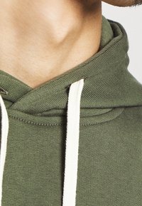Sudadera color verde oliva con tejido texturizado, que incluye un cordón, un cuello acanalado y un ajuste relajado. Primer plano de la zona del cuello.