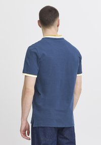 Temno moderna polo majica z rumeno in belo prugasto ovratnikom, kratkimi rokavi in stranskimi razporki, ki prikazuje gladko bombažno teksturo.