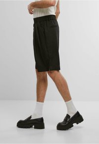 Svarte skreddersydde shorts med bred elastisk midjebånd, sidelommer og løs passform. Bærer hvite ribbede sokker og kraftige svarte sko.