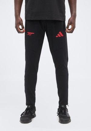 Mand iført sorte sportbukser med røde Adidas- og Arsenal-logoer står med hænderne nær hofterne mod en hvid baggrund.