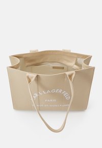 Beige kangaskassi, jossa on kaksinkertaiset kahvat, suuri valkoinen logo ja teksti. Sisus on vuoraamaton, ja siinä on yksi vetoketjullinen osasto.