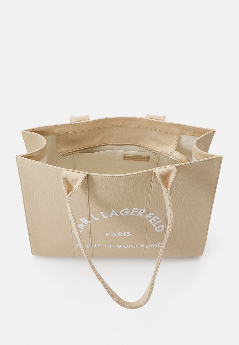 Beige kangaskassi, jossa on kaksinkertaiset kahvat, suuri valkoinen logo ja teksti. Sisus on vuoraamaton, ja siinä on yksi vetoketjullinen osasto.