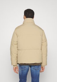 Beige puffer jacket with high collar, quilted design, and padded texture. Worn over a dark sweater, complemented by blue jeans.