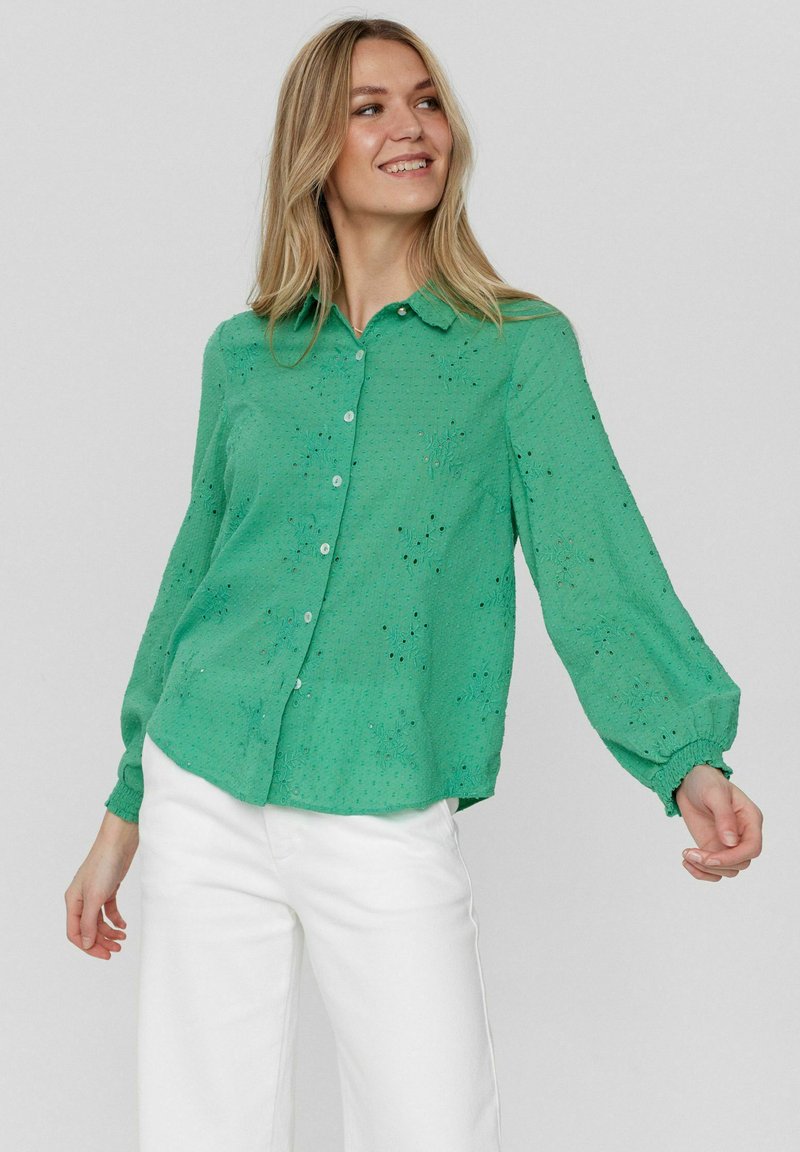 Camicia verde con bottoni, motivo floreale ricamato, maniche lunghe a palloncino e orlo arrotondato, abbinata a pantaloni bianchi.