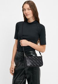 Donna con lunghi capelli castani che indossa un top nero a maniche corte e pantaloni neri lucidi, mentre tiene in mano una borsa a tracolla nera con un motivo testuale bianco.