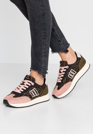 Pieds portant un jean noir et des baskets roses, marron et noires avec un imprimé animalier et un logo blanc, debout sur un sol gris clair.