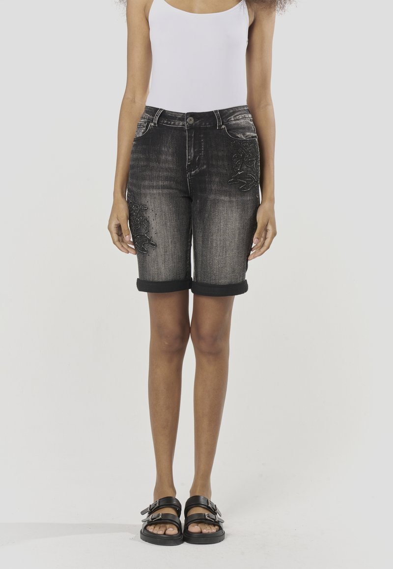 Schwarze Jeansshorts mit umgeschlagenen Säumen, mit floraler Stickerei am linken Bein. Kombiniert mit schwarzen Sandalen und einem weißen Tanktop.