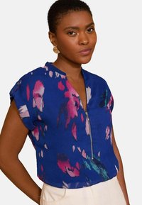 Top azzurro a maniche corte con zip parziale e motivi floreali rosa. Tessuto leggero con una consistenza liscia. Vestibilità casual.