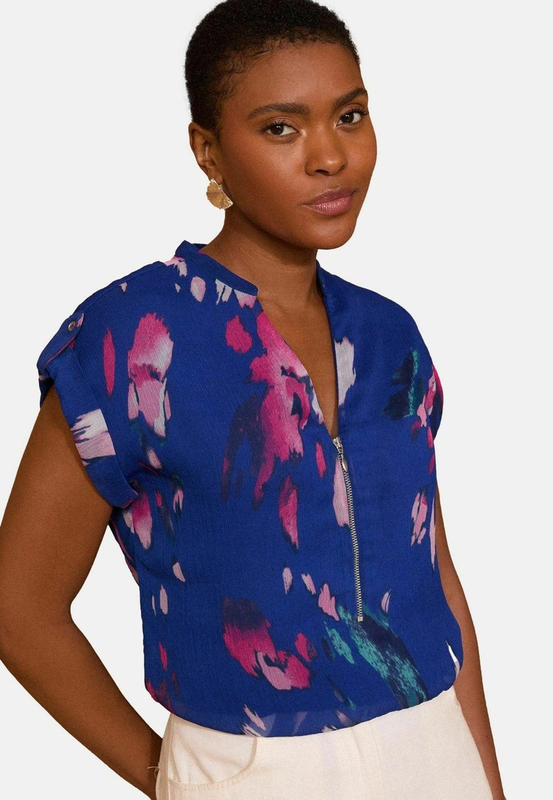 Top azzurro a maniche corte con zip parziale e motivi floreali rosa. Tessuto leggero con una consistenza liscia. Vestibilità casual.