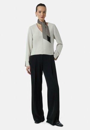 Lichtgrijs langemouwen top met een V-hals en een zwart-wit gestreepte sjaal om de nek. Zwarte wijde broek en platte zwarte schoenen maken de outfit compleet.