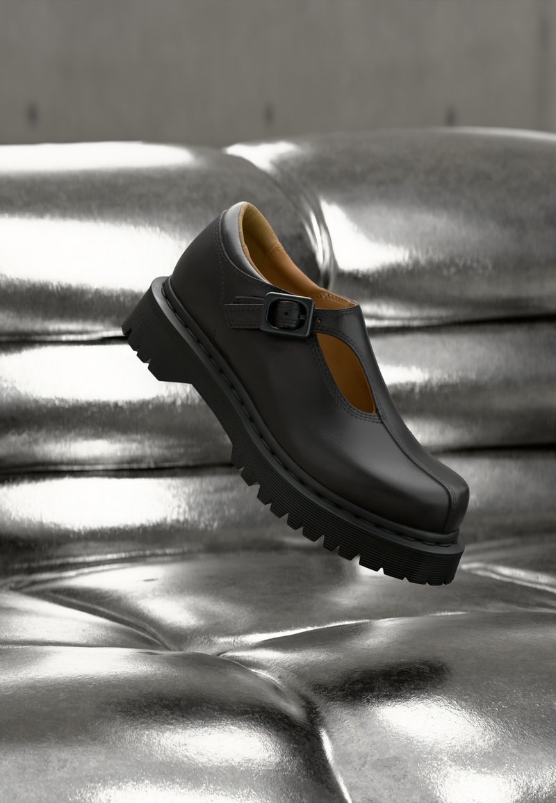 Scarpa Mary Jane in pelle nera con suola spessa e scanalata e cinturino con fibbia, sospesa sopra una superficie argentata lucida e capitonnè.