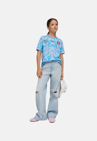 T-shirt graphique bleu clair avec motifs abstraits et drapeau suisse, associé à un jean déchiré bleu clair et des chaussures roses. Casquette blanche tenue à la main.