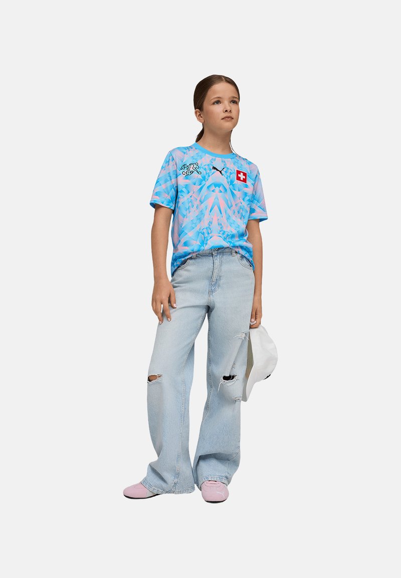 T-shirt graphique bleu clair avec motifs abstraits et drapeau suisse, associé à un jean déchiré bleu clair et des chaussures roses. Casquette blanche tenue à la main.