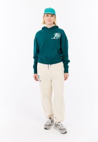 Tyrkysová mikina s klokaní kapsou a grafickým logom, skombinovaná s krémovými nohavicami s potlačou loga a sivými teniskami.