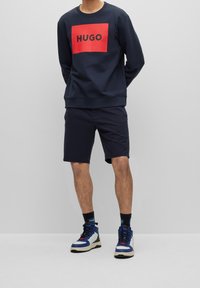 Sudadera navy con el logotipo en rojo "HUGO", combinada con pantalones cortos navy y zapatillas altas azules. Estilo simple, corte relajado con tejido texturizado.
