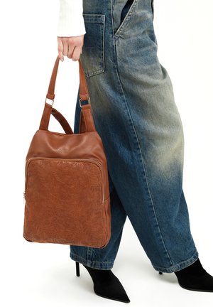 Bolso de cuero marrón con grabado floral, un bolsillo frontal con cremallera, doble correa y forma rectangular. Llevado con pantalones de mezclilla anchos azules.