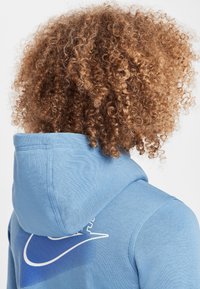 Light blue hoodie with a soft texture, featuring a large white Nike logo on the back and a drawstring hood. Curly brown hair is visible.