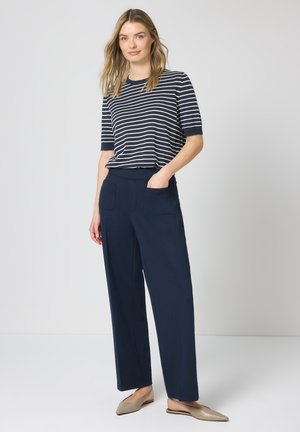 Kvinde står med hånden i lommen iført navyblå kortærmet top med striber, navyblå bukser med vide ben og beige spidse flade sko mod hvid baggrund.