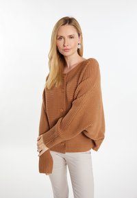 Bruine geribde cardigan met oversized mouwen en knoopsluiting. Soepele textuur met een ontspannen pasvorm, gedragen over lichtgekleurde broeken.