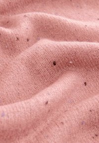 Měkká růžová pletenina s texturovaným, jemným vzorem. Obsahuje malé tmavé a světlé skvrny, čímž vytváří vizuální zajímavost a hloubku.
