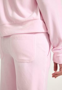 Różowy, miękki dres o swobodnym kroju. Spodnie mają widoczną tylną kieszeń, a przeszycia podkreślają gładką fakturę materiału.